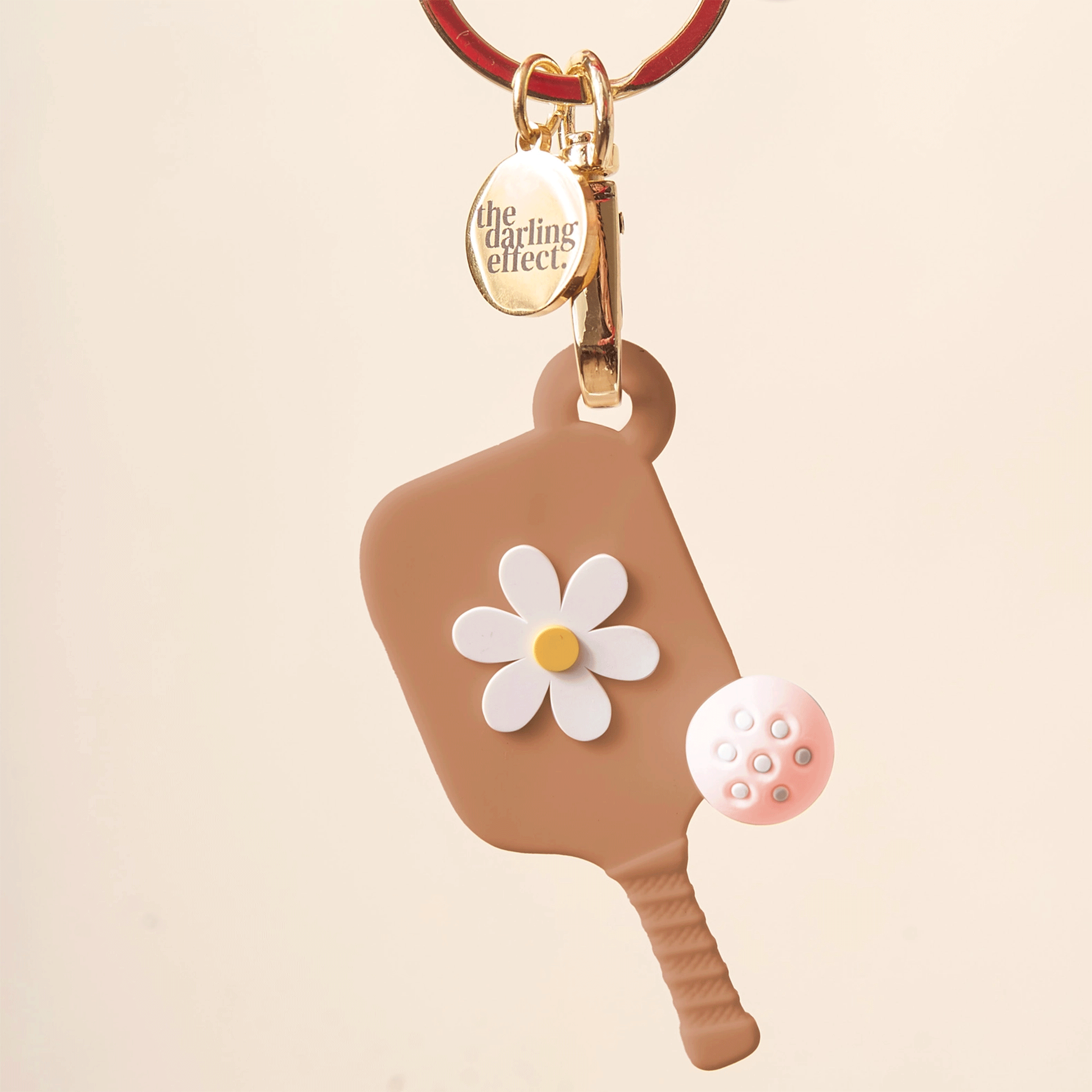 On an ivory background is a keychain with a tan pickleball paddle charm with a daisy in the middle. 