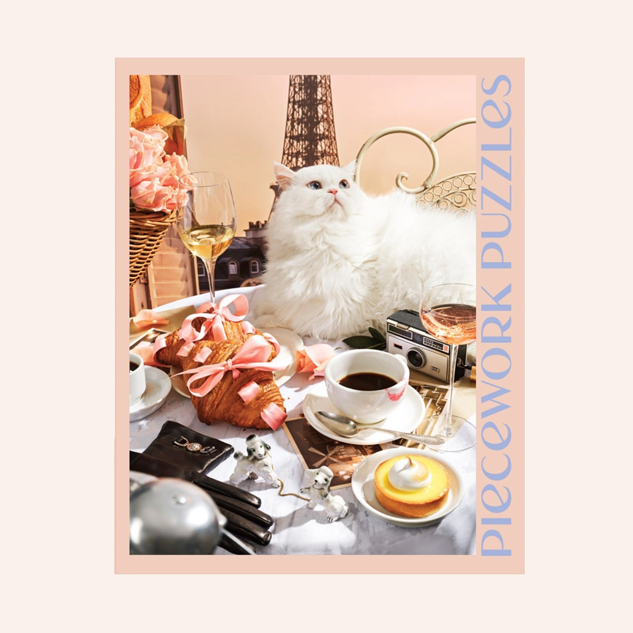 A peachy tan colored puzzle box with a 1000 piece puzzle inside with an image of a white fluffy cat surrounded by French goods such as croissants, macarons, champagne and the Eiffel Tower. 