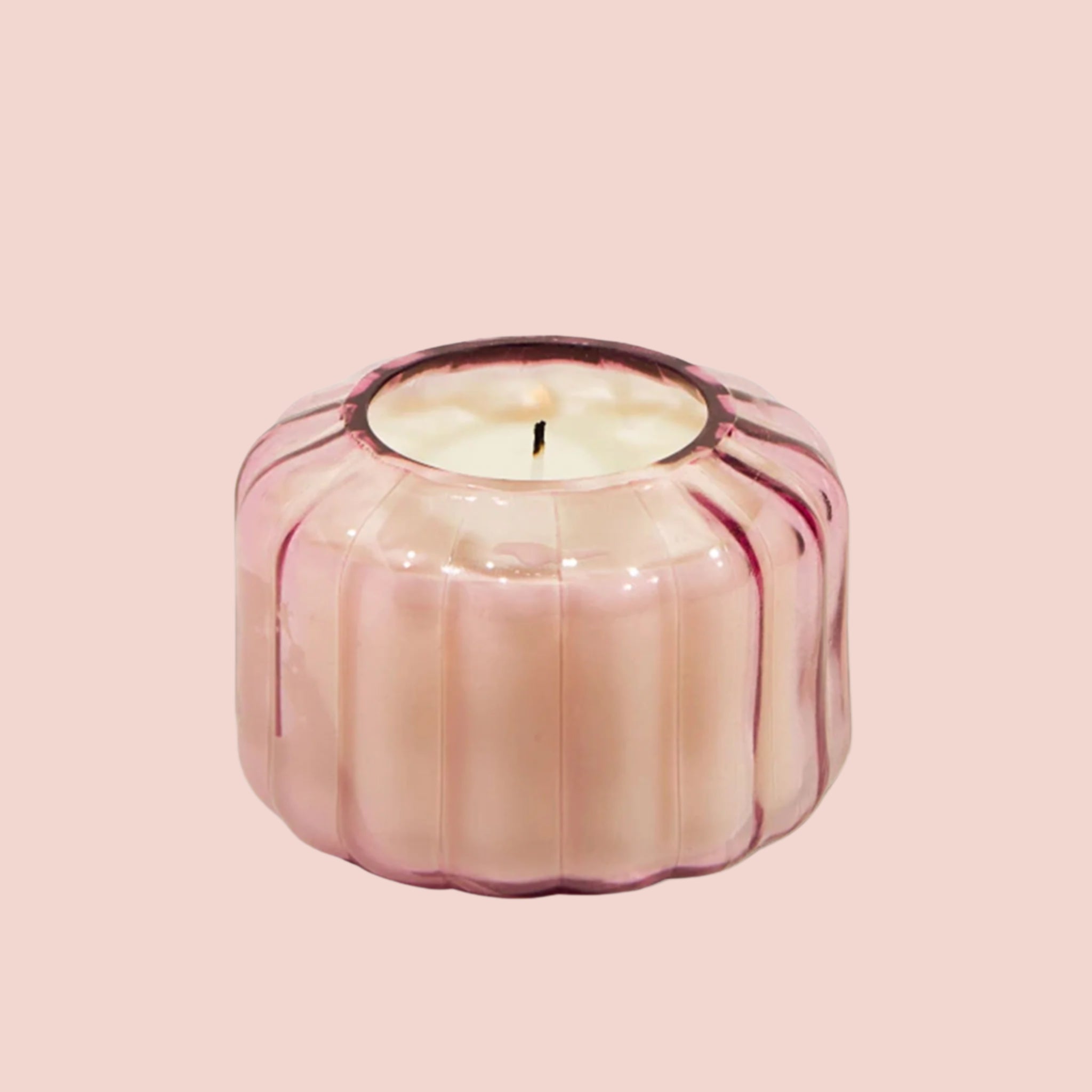 On a pink background is a pink glass candle with a ribbed texture and a single wick.