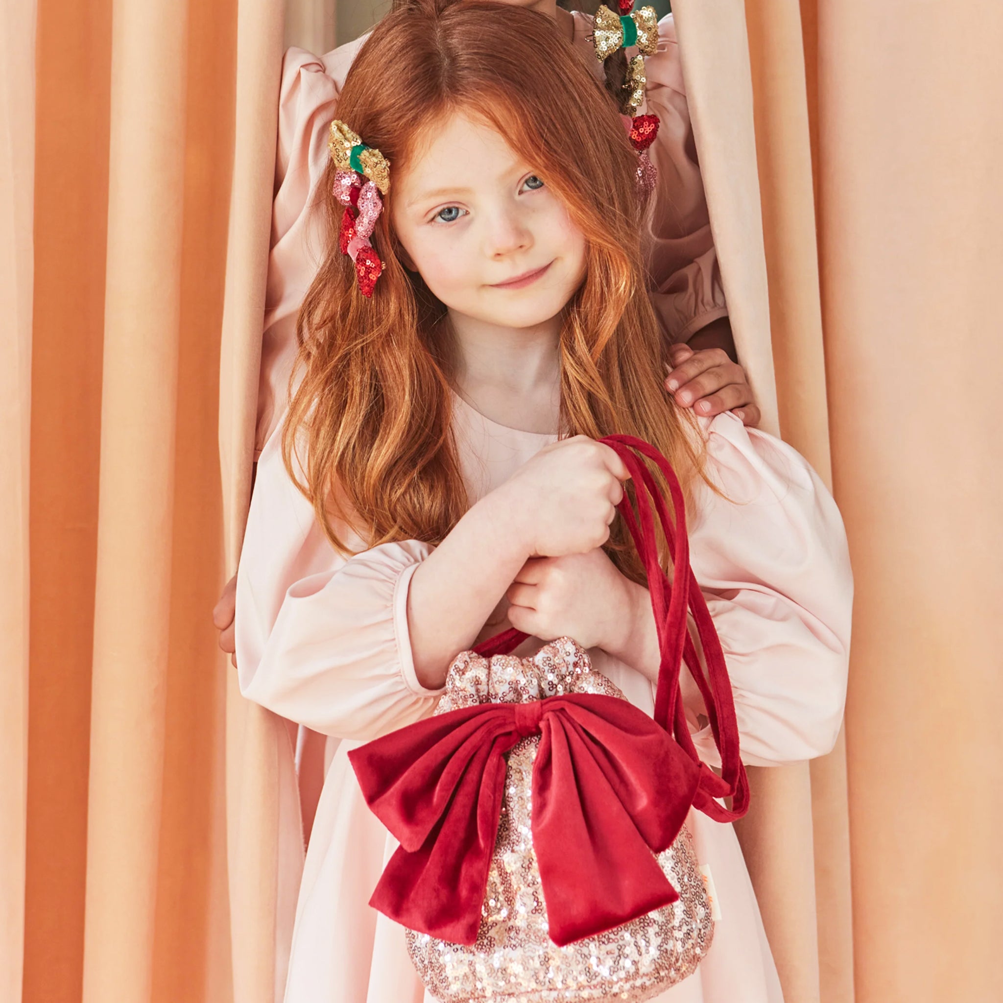 A dusty pink sequin pouch bag with a red velvet bow and strap. 