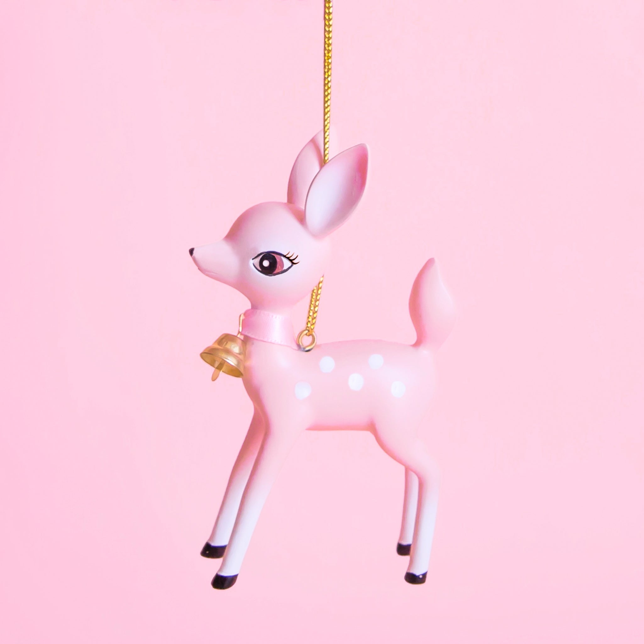 On a pink background is a light pink retro deer ornament with white details and spots and a gold bell on the front of its neck along with a gold string loop for hanging.