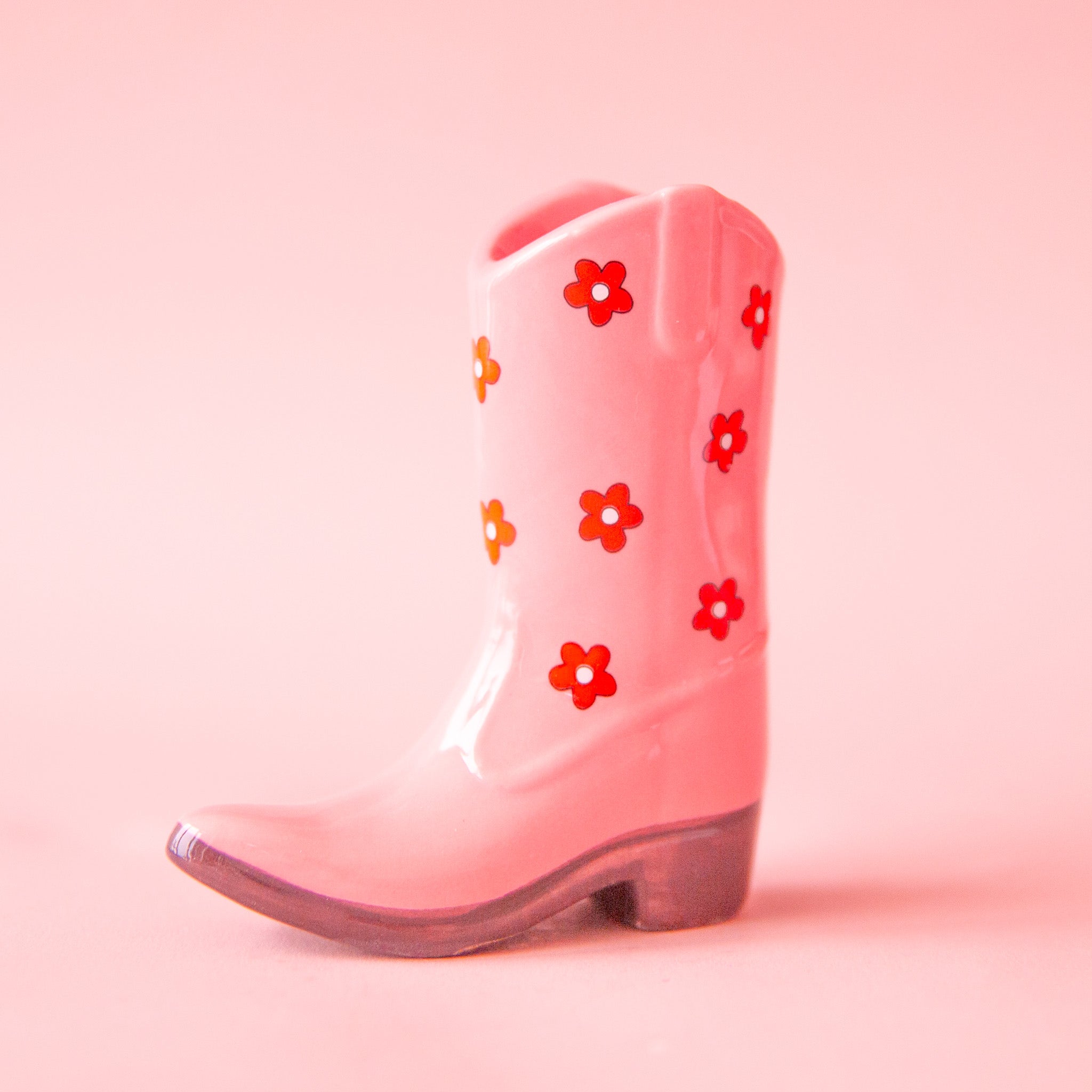 On a pink background is a pink cowboy boot shaped vase with a red and white daisy print on it.