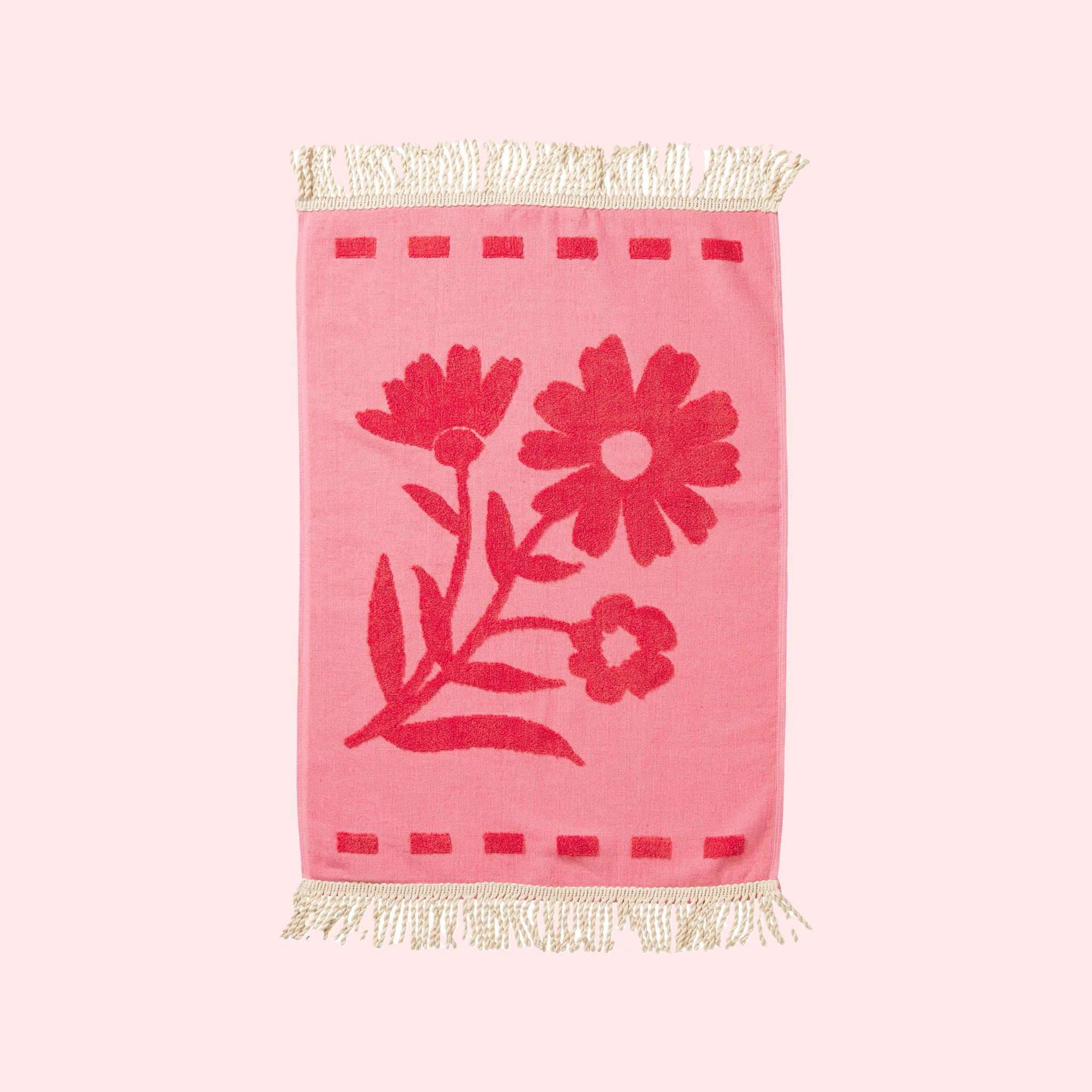 A light and dark pink hand towel with a floral pattern and fringe detailing on two ends. 