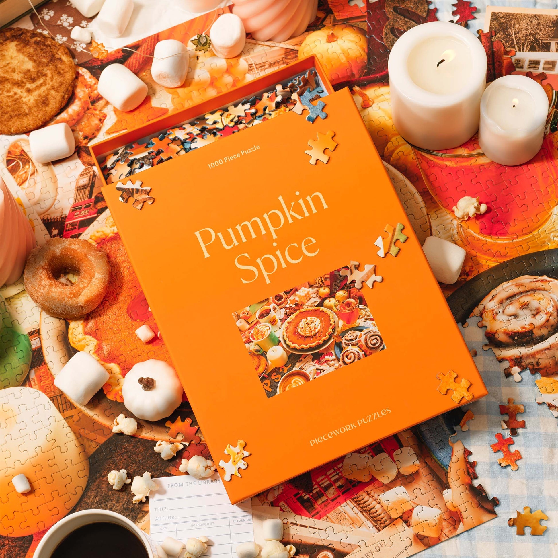 An orange puzzle box that reads &quot;Pumpkin Spice&quot; site on top of a half-worked puuzzle below.  In the backround are lit white candles, popcorn sprinkled about, marshmallows sprinkled about, an old fashioned donut and a cup of cofee.  The image evokes the feeling of Fall colors and mood.