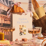 A peachy tan colored puzzle box with a 1000 piece puzzle inside with an image of a white fluffy cat surrounded by French goods such as croissants, macarons, champagne and the Eiffel Tower. 