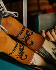 A model wearing a pair of burnt orange crew socks with a black scorpion graphic and text that reads, "Quesadilla".
