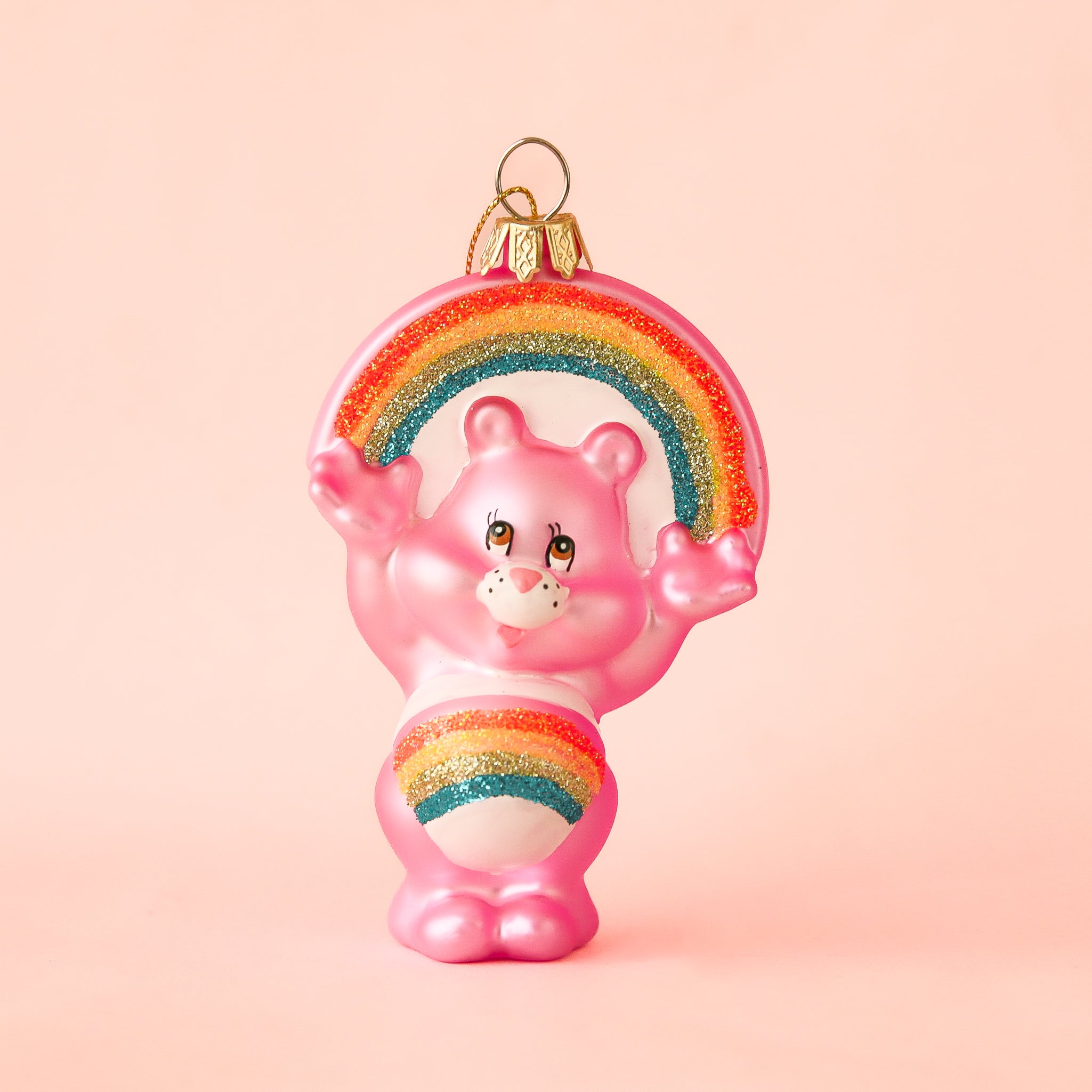 On a peachy background is a pink glass bear shaped ornament with a rainbow on its stomach and arched over its head. 