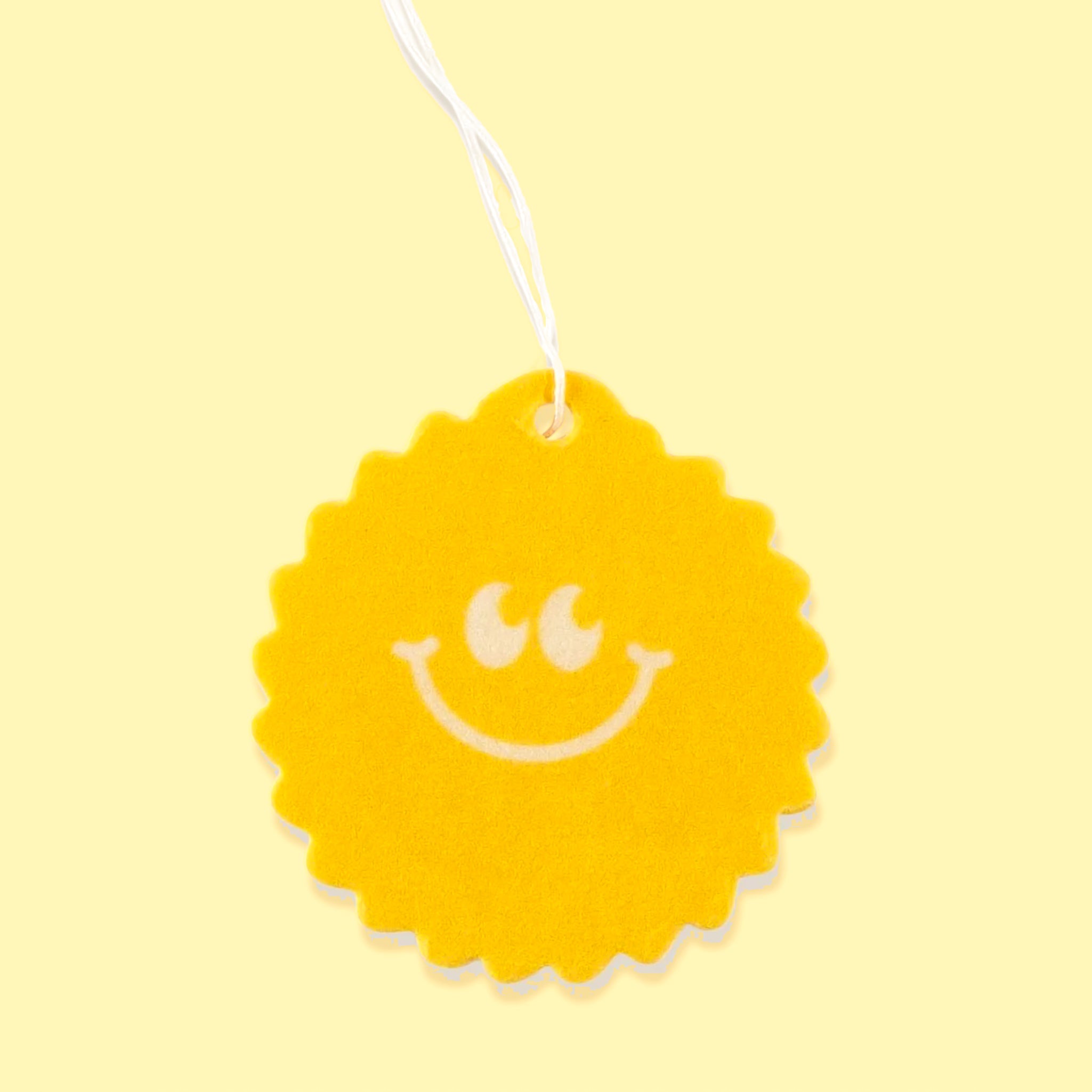 On a yellow background is a yellow sunshine shaped air freshener with a smiley face and a white hanging string. 