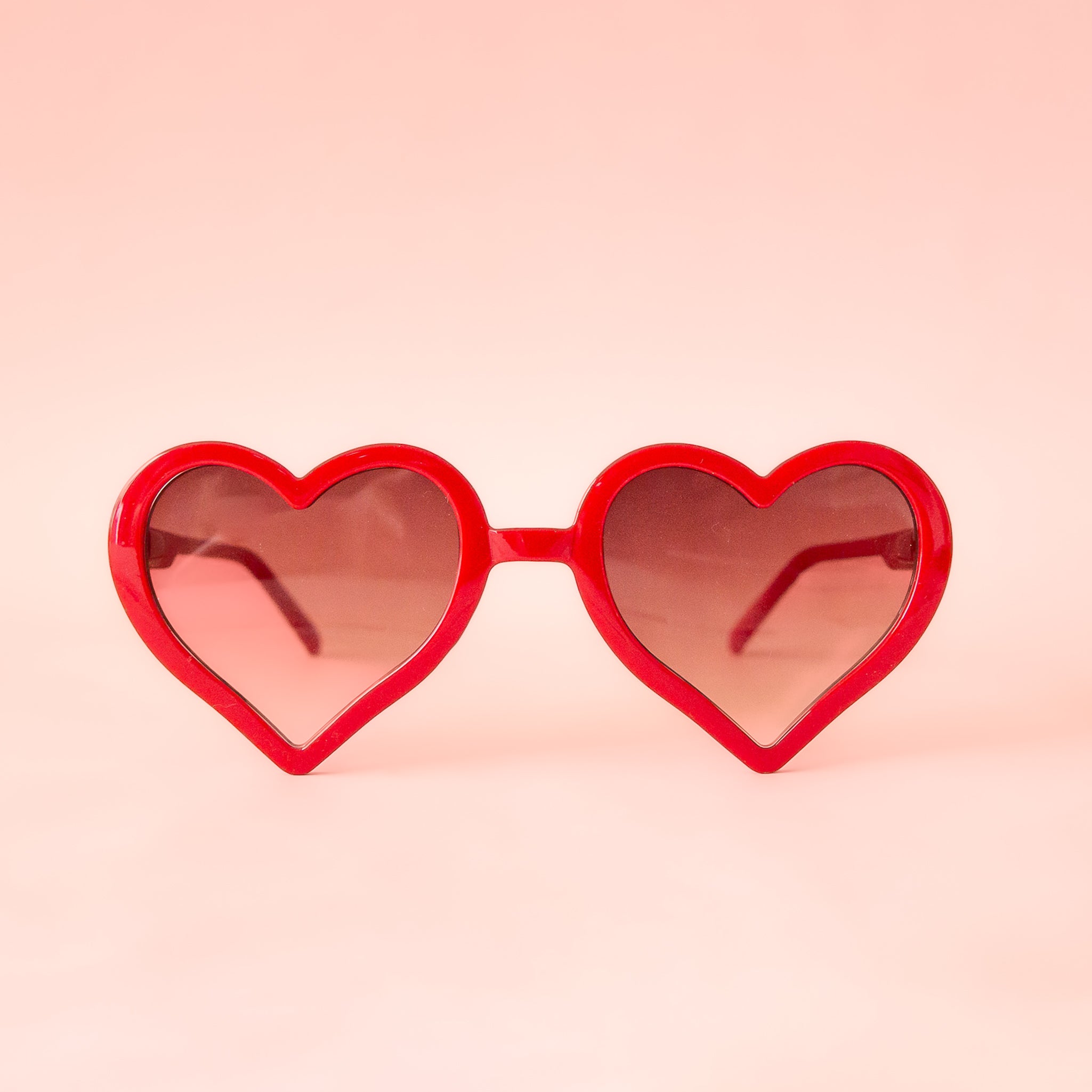 On a pink background is a red pair of heart shaped sunglasses.