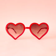 On a pink background is a red pair of heart shaped sunglasses.