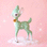 On a pink background is a mint green retro deer figurine with white spot details and a gold bell at the front of its neck.