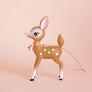 On a peachy background is a brown retro deer ornament with white details and spots and a gold bell on the front of its neck along with a gold string loop for hanging.