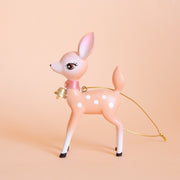 On a peach background is a peachy pink retro deer ornament with white details and spots and a gold bell on the front of its neck along with a gold string loop for hanging.