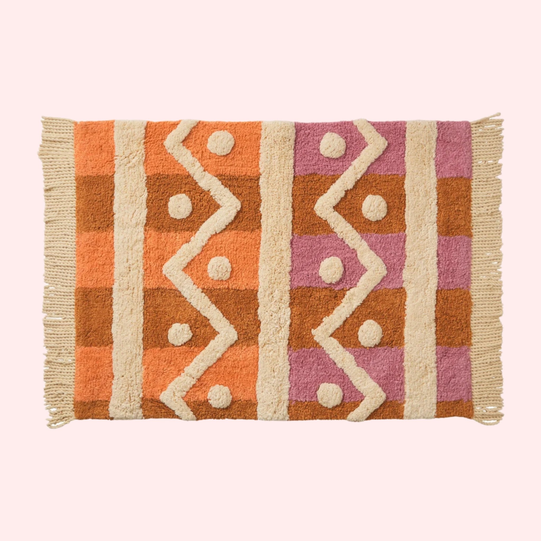 A pink, orange and ivory tufted bath mat with a geometric pattern and tassel details on two ends. 