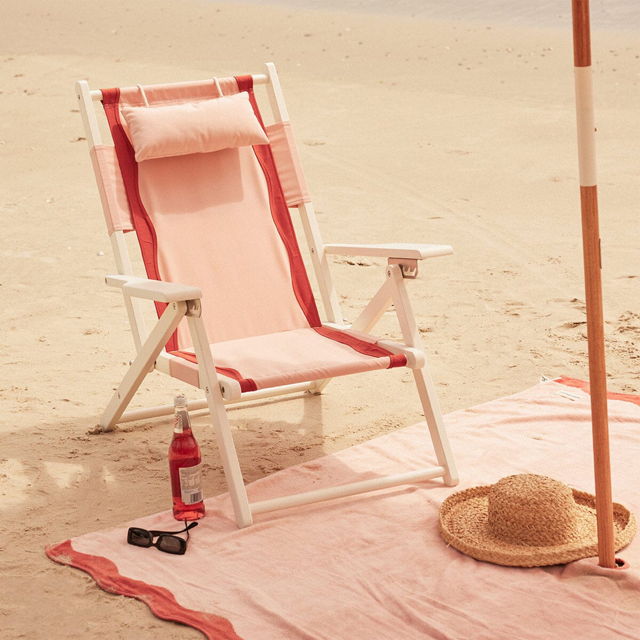 A pink and white folding beach chair with dark pink / reddish accents, a head support pillow and a zip pocket on the back of the chair. 