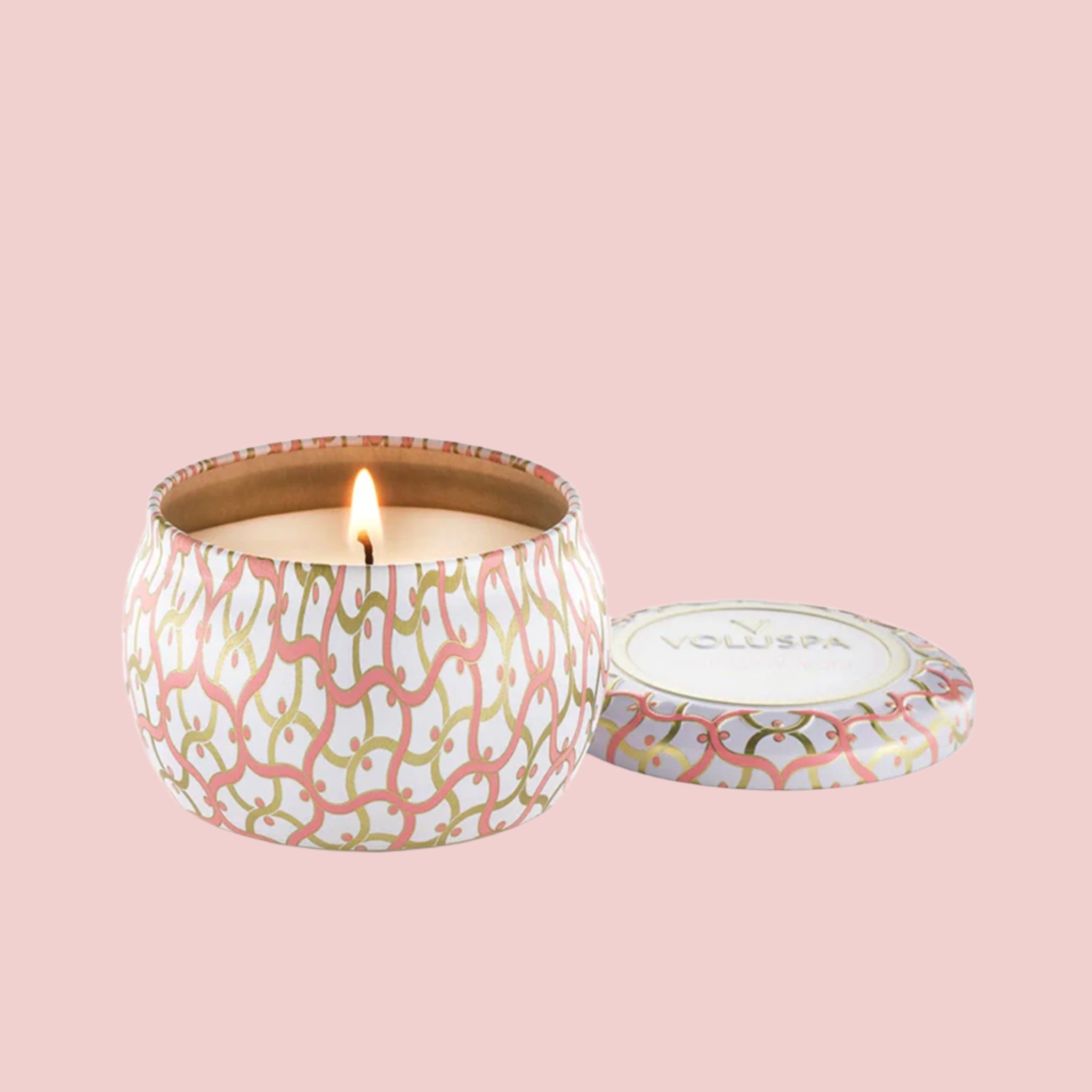 A round tin candle. The tin is white with pink and gold swirly lines. Inside the tin is a white candle with a white wick in the center. There is a matching lid leaning against the left side of the candle.