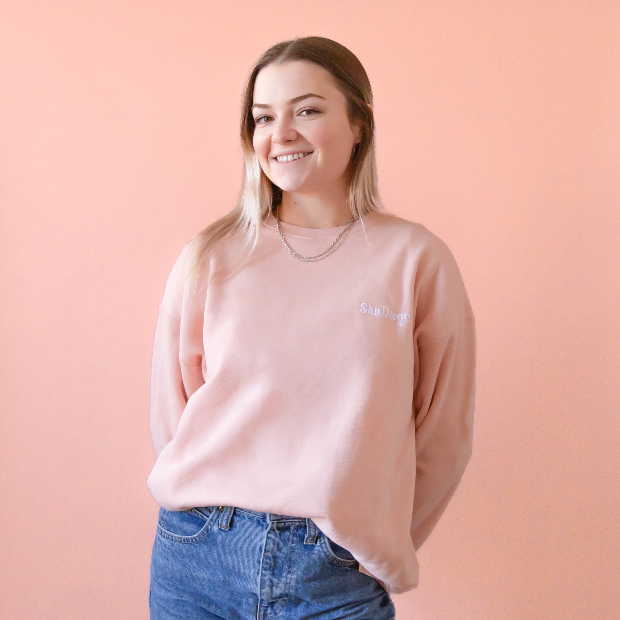 On a peach background is a peachy pink pull over sweatshirt with a white embroidered "San Diego" on the front.