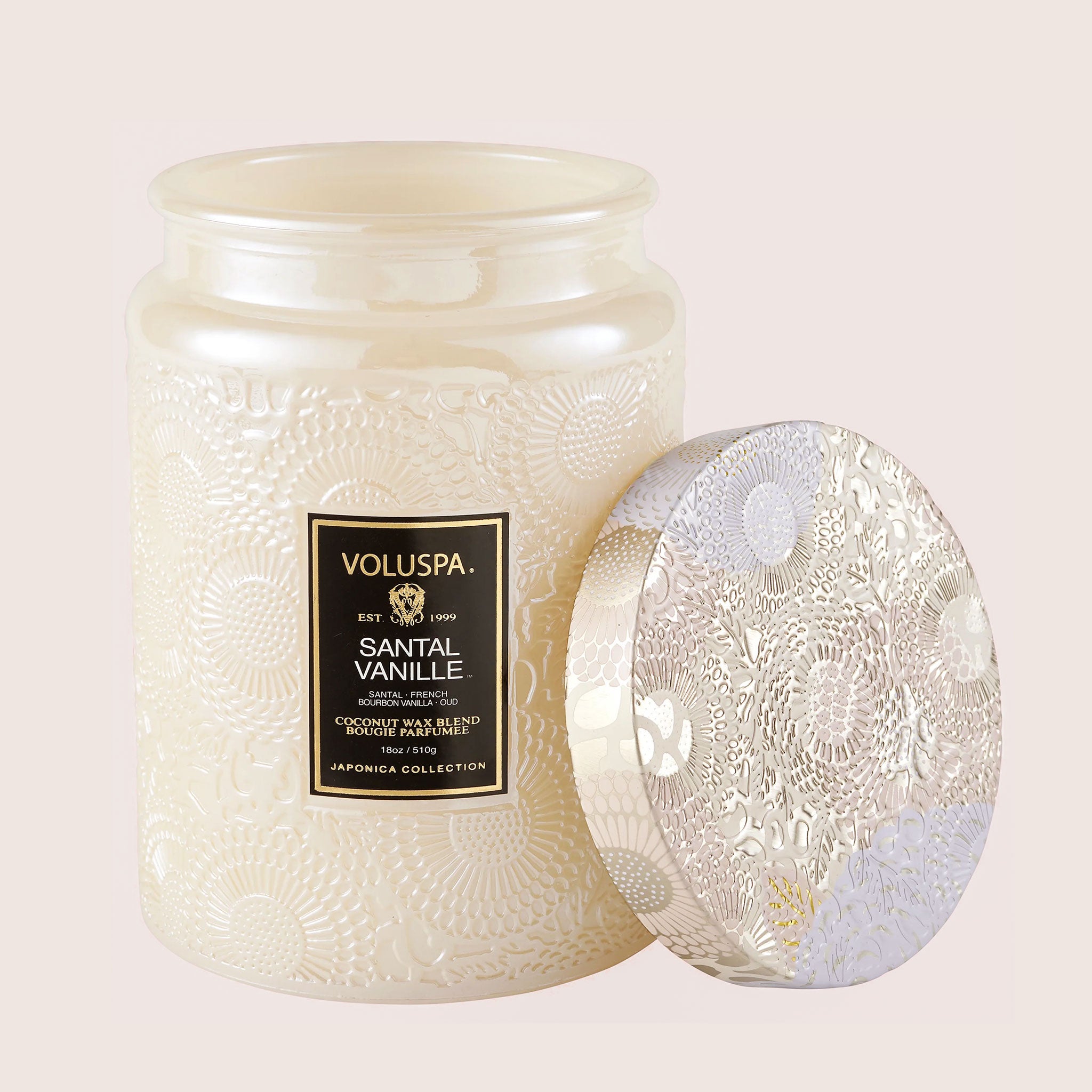 In front of a white background is a large light pink glass jar. The glass jar has a floral pattern all over it. In the middle is a black rectangular sticker with gold text at the top that reads ‘voluspa.’ Below is white text that reads ‘santal vanille.’ Leaning against the right side of the jar is a round tin lid. The lid has a light pink, silver and lavender flower pattern on it.