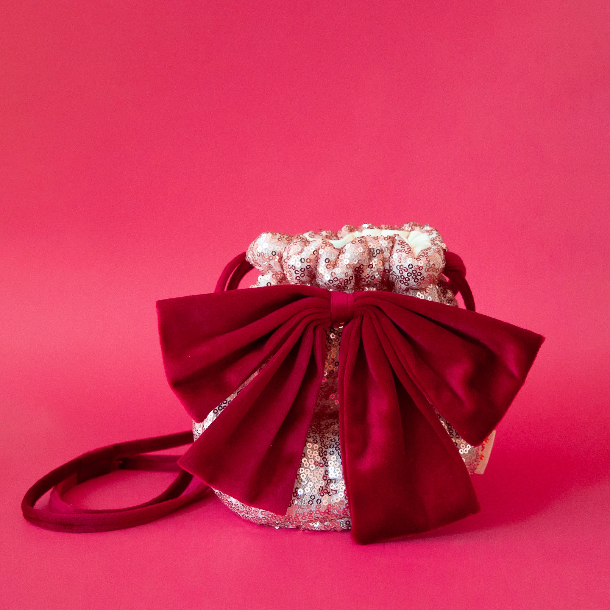 A dusty pink sequin pouch bag with a red velvet bow and strap. 