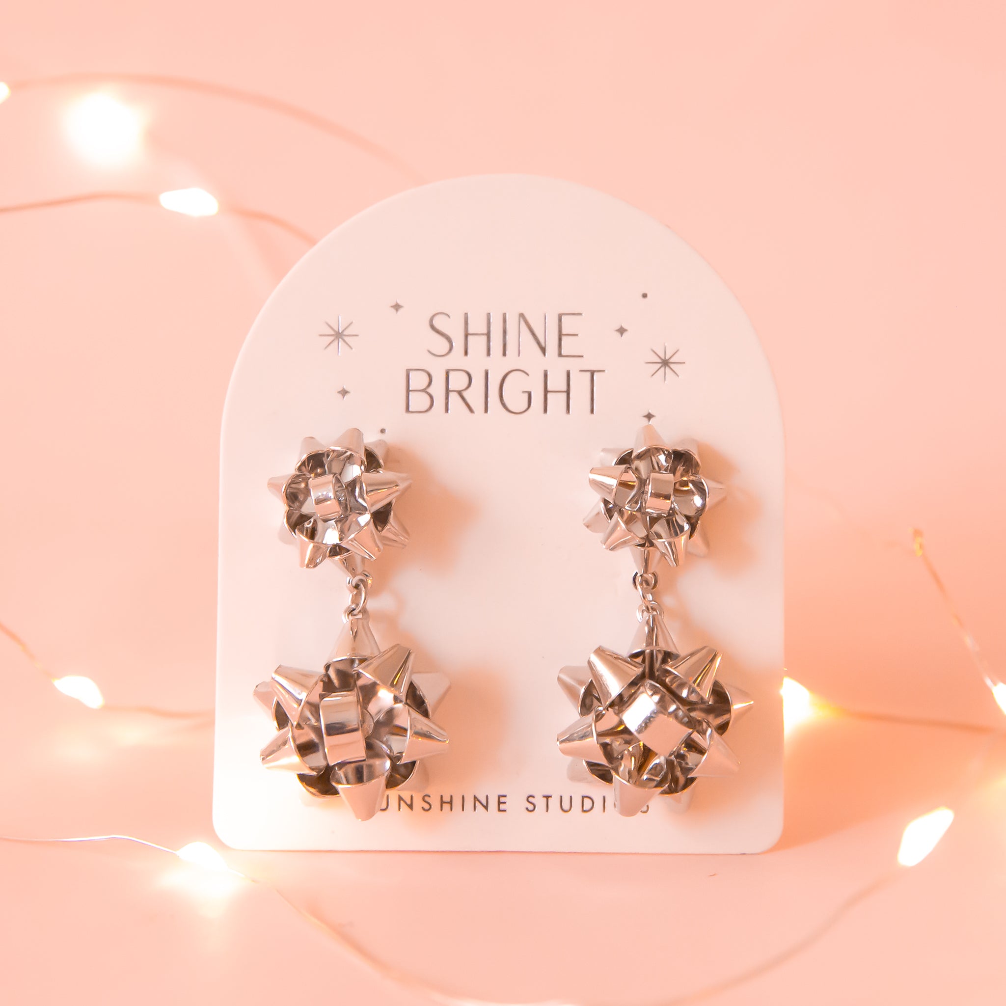 A pair of silver holiday bow shaped earrings that have a smaller gold bow and a dangling larger bow attached to the bottom. The packaging reads, &quot;Shine Bright Sunshine Studios&quot;.