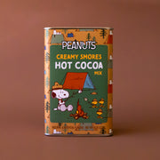 A green and orange metal tin with a camping Snoopy design and text that reads, 'Peanuts Creamy Smores Hot Cocoa Mix'. 