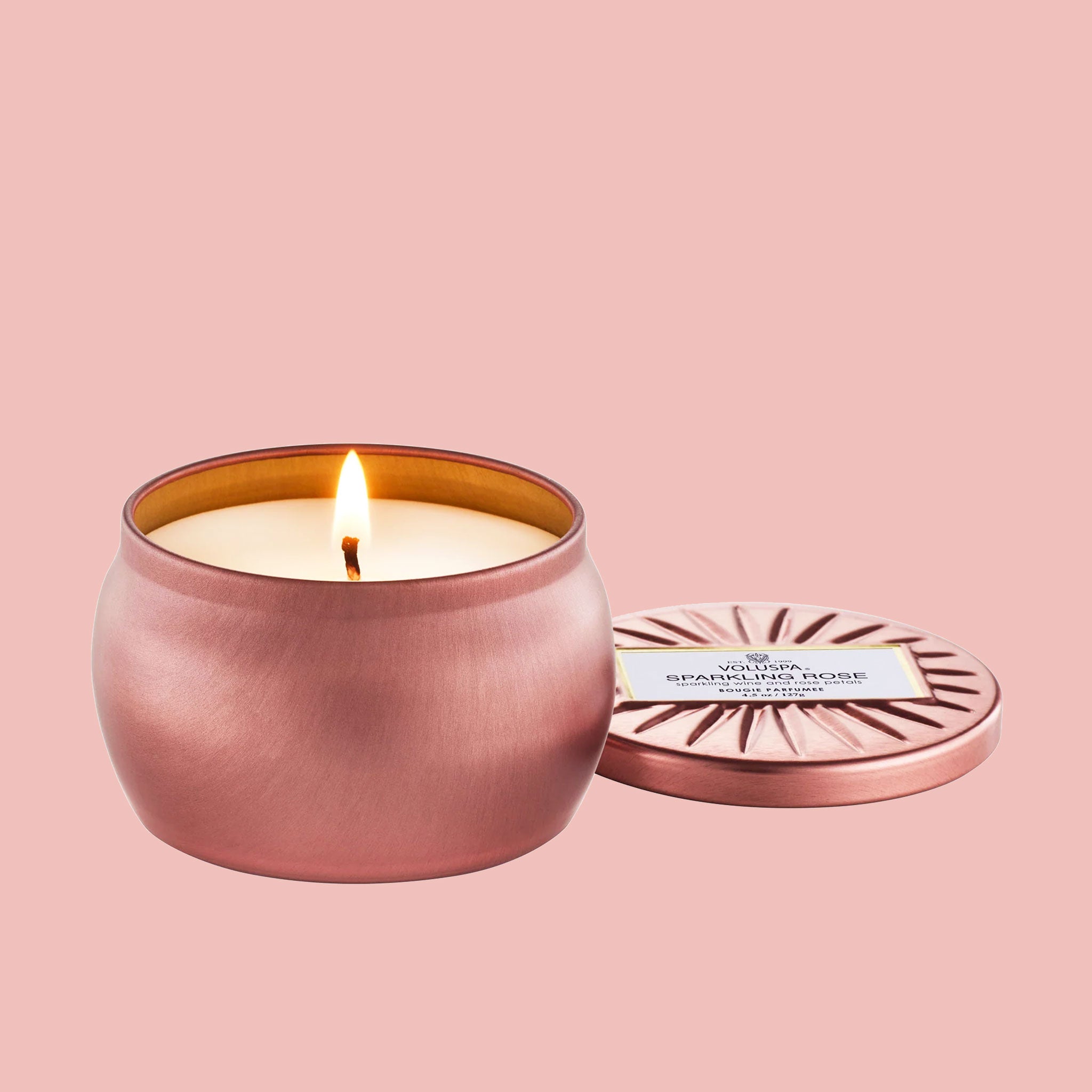 A rosy pink round candle sits in the middle of frame with the lid off to the right.  The tin is a metallic pink color and the candle flame is lit on open candle side.  The lid sits to the right of the candle and reads &quot;Voluspa Sparkling Rose&quot;.