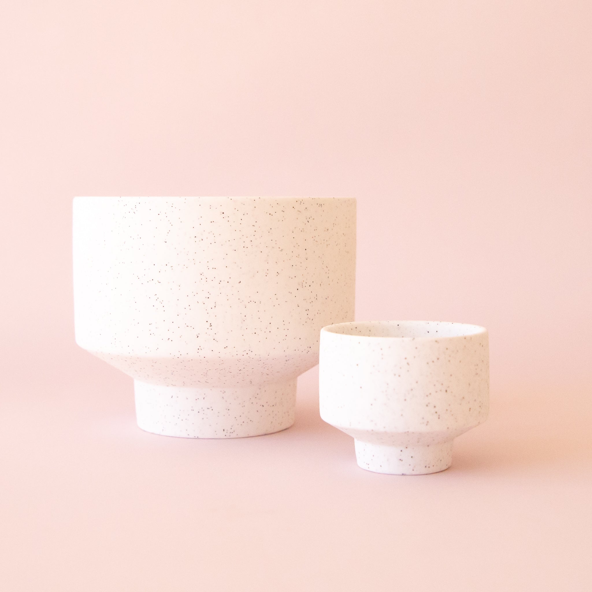 A speckled footed ceramic planter in a neutral shade next to the larger version. 