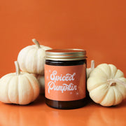 Against an orange background is a dark amber glass candle jar with an orange label that reads, "Spiced Pumpkin" in white cursive font. Also photographed are 5 white pumpkins staged beside the candle. Pumpkins not included.