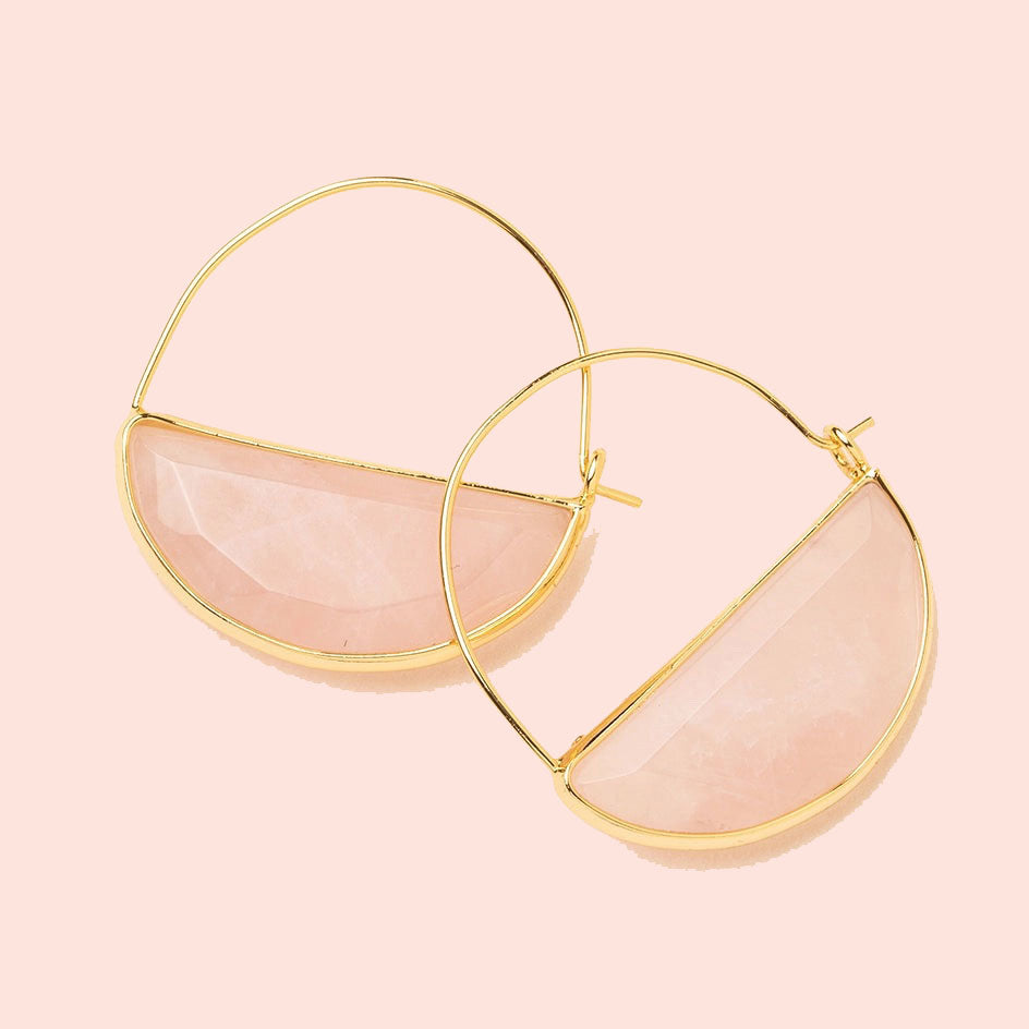 In front of a cream background is a pair of gold earrings. The top is a thin gold hoop. The bottom is a rose quartz half circle with a gold border. 