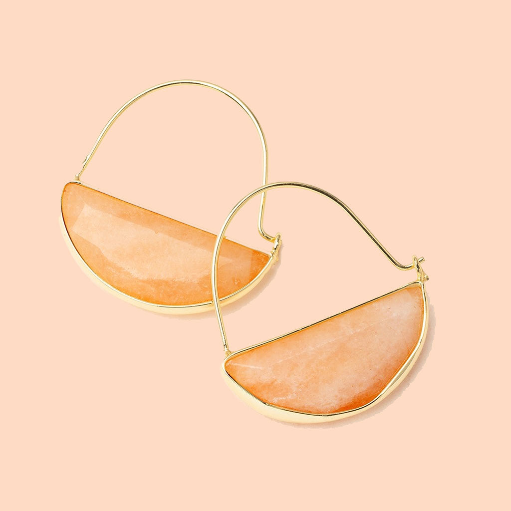 In front of a cream background is a pair of gold earrings. The top is a thin gold hoop. The bottom is a peach stone half circle with a gold border. 