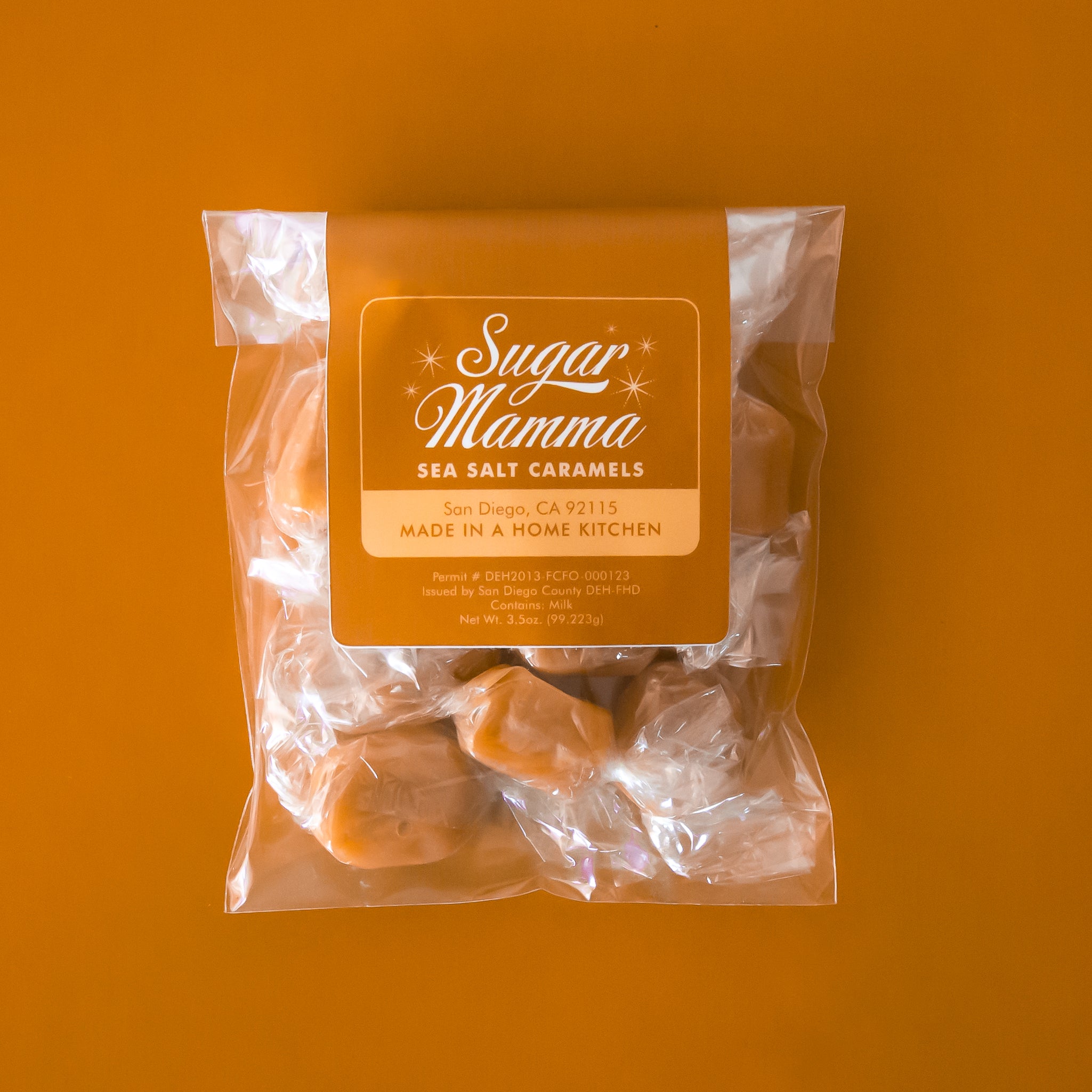 This is a clear, square plastic package. Inside the package is light brown candies. There is a light orange vertical belly band that stops before touching the bottom. In the middle of the belly band is a white outlined square. Inside is white script that reads ‘sugar mamma.’ Below is more white text that reads ‘sea salt caramels.’ The bottom half of the square is peach with light orange text that reads ‘San Diego, CA 92115 Made in a home kitchen.’ 