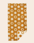 A cream and mustard yellow beach / bath towel with a sun design on the front and back, photographed here with the mustard side facing up and a corner of the towel flipped over to show the opposite cream side. 