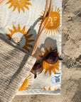 A cream and mustard yellow beach / bath towel with a sun design on the front and back and photographed on a sand beach alongside a pair of sunglasses and a woven straw bag.  