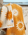 A cream and mustard yellow beach / bath towel with a sun design on the front and back, photographed here draped over the edge of a concrete barrier in front of the beach. 