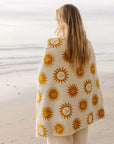 A cream and mustard yellow beach / bath towel with a sun design on the front and back wrapped around a model's shoulders in this photo. 