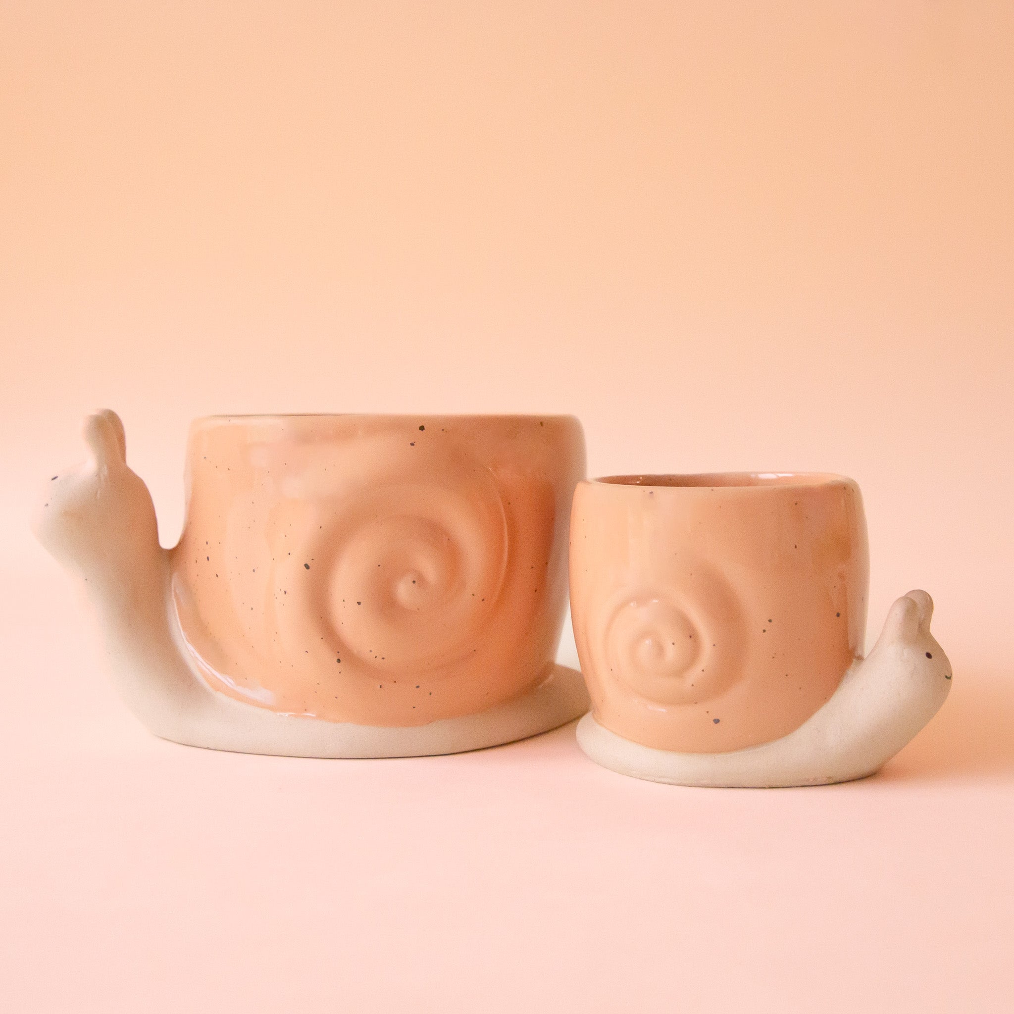 On a light peach background is two ceramic planters in the shape of a snail with a swirly light orange "shell". 