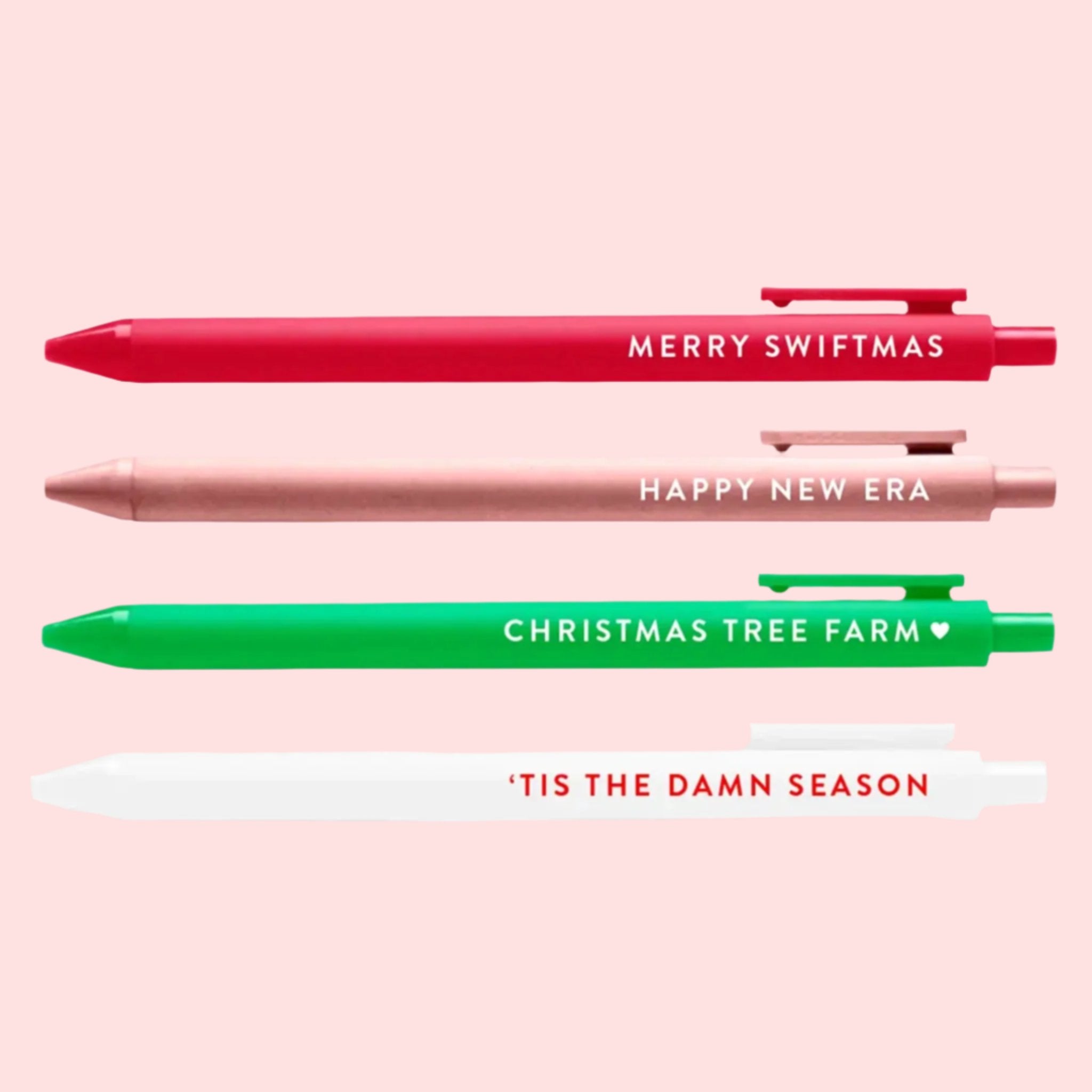 A pack of four black ink pens in red, pink, green and white with text on each that reads a different Taylor Swift related phrase. From top to bottom the phrases reads, &#39;Merry Swiftmas&#39;, &#39;Happy New Era&#39;, &#39;Christmas Tree Farm&#39; and &#39;Tis The Damn Season&#39;. 