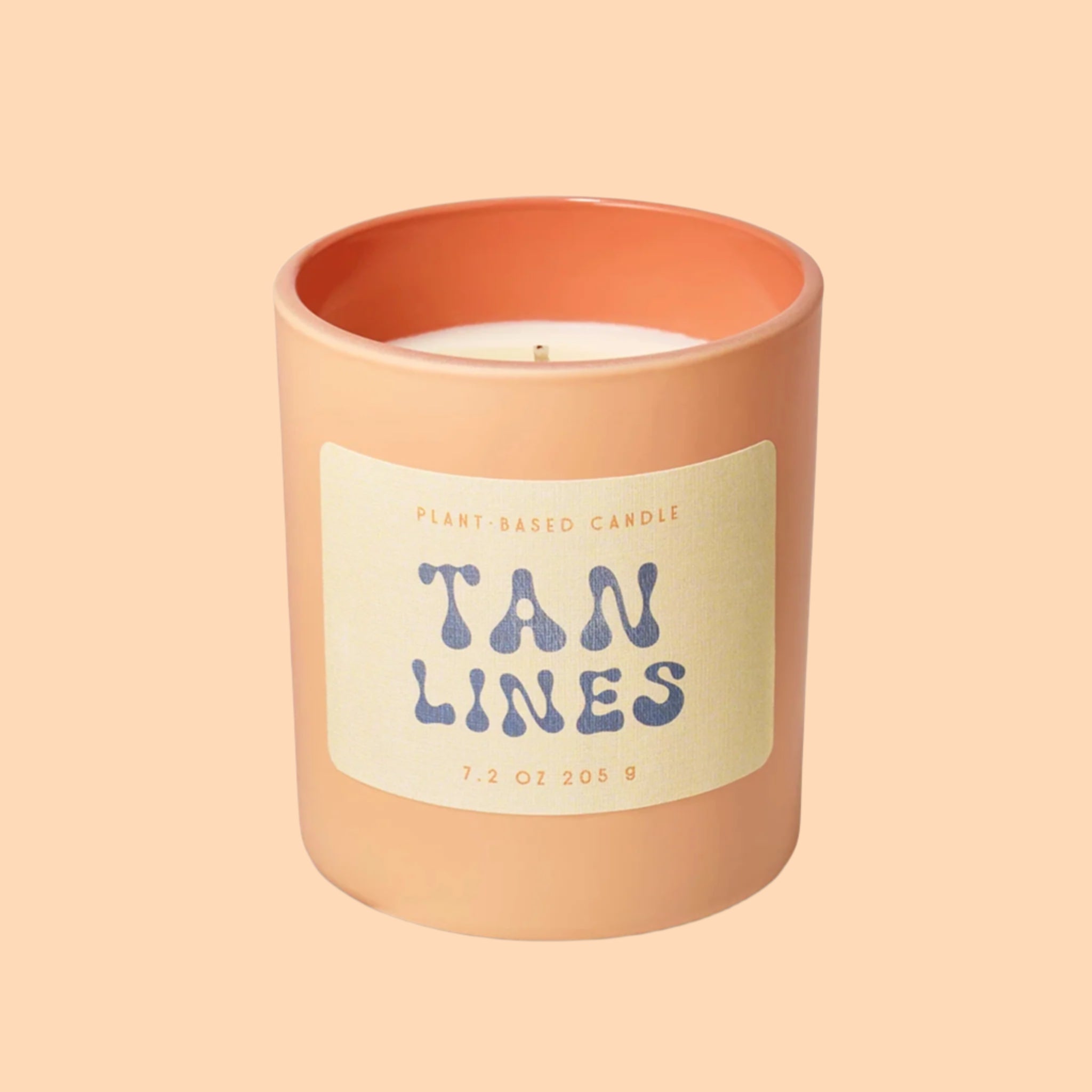 An apricot colored glass candle with a tan label, blue text that reads, 'Tan Lines'. 