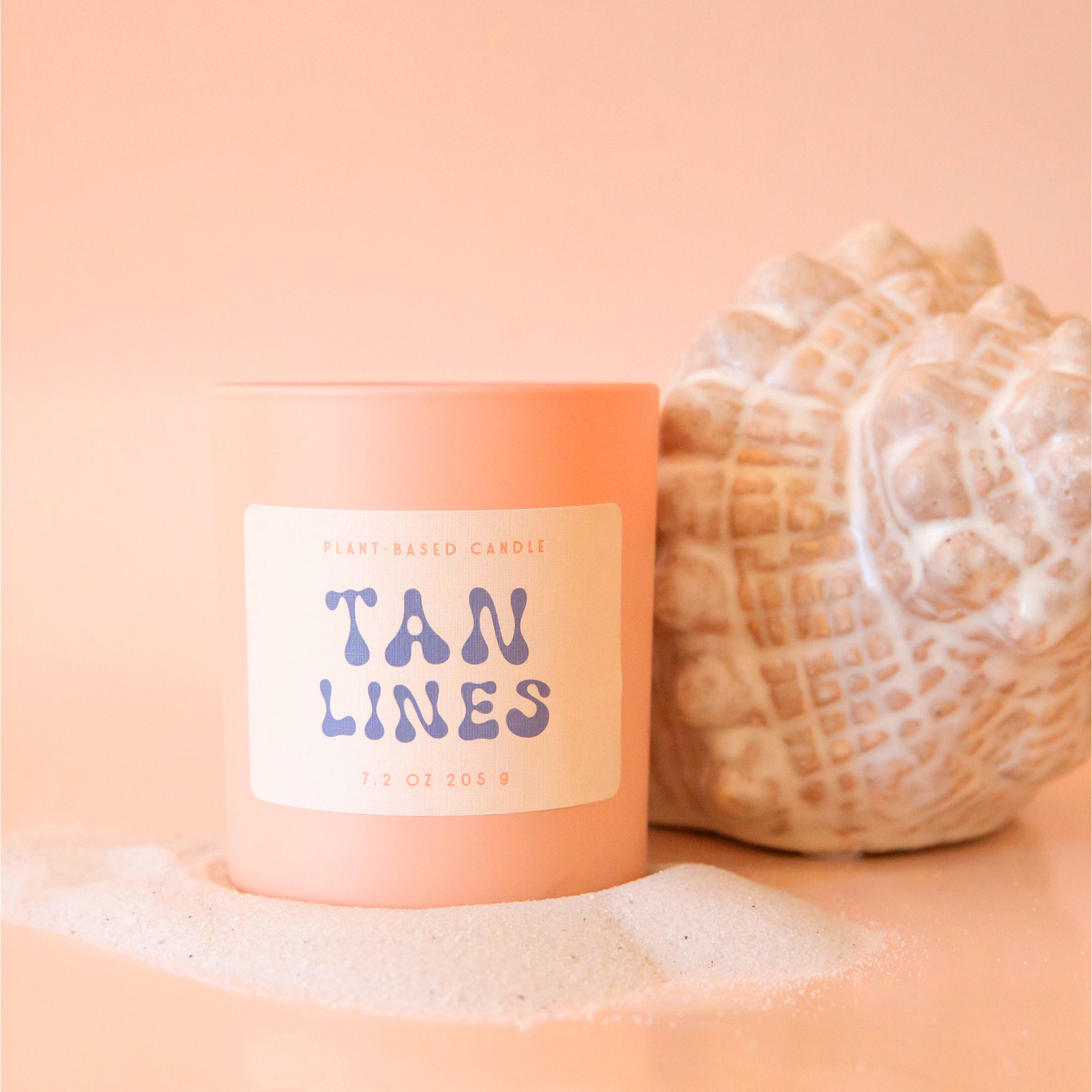 An  apricot colored glass candle with a tan label, blue text that reads, 'Tan Lines'. 