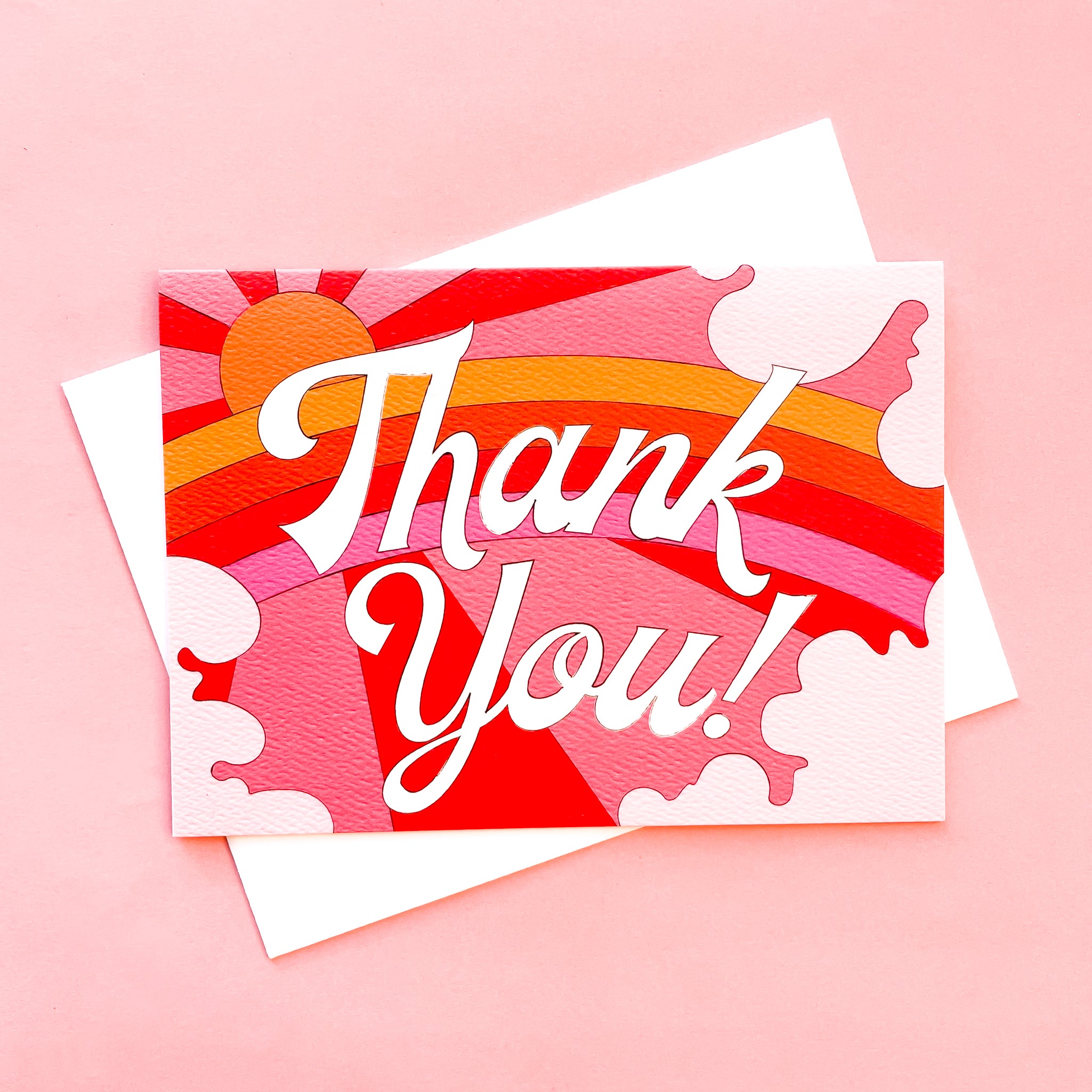 Card with a vibrant scene of a rosie toned rainbow and beaming pink and red sun. Soft pink cloud take up the edges. The center of the card reads 'Thank you!' in white cursive lettering.