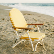 A yellow arched back folding beach chair with wood armrests and white legs. The backside features a zip pocket for storing beachside essentials. 