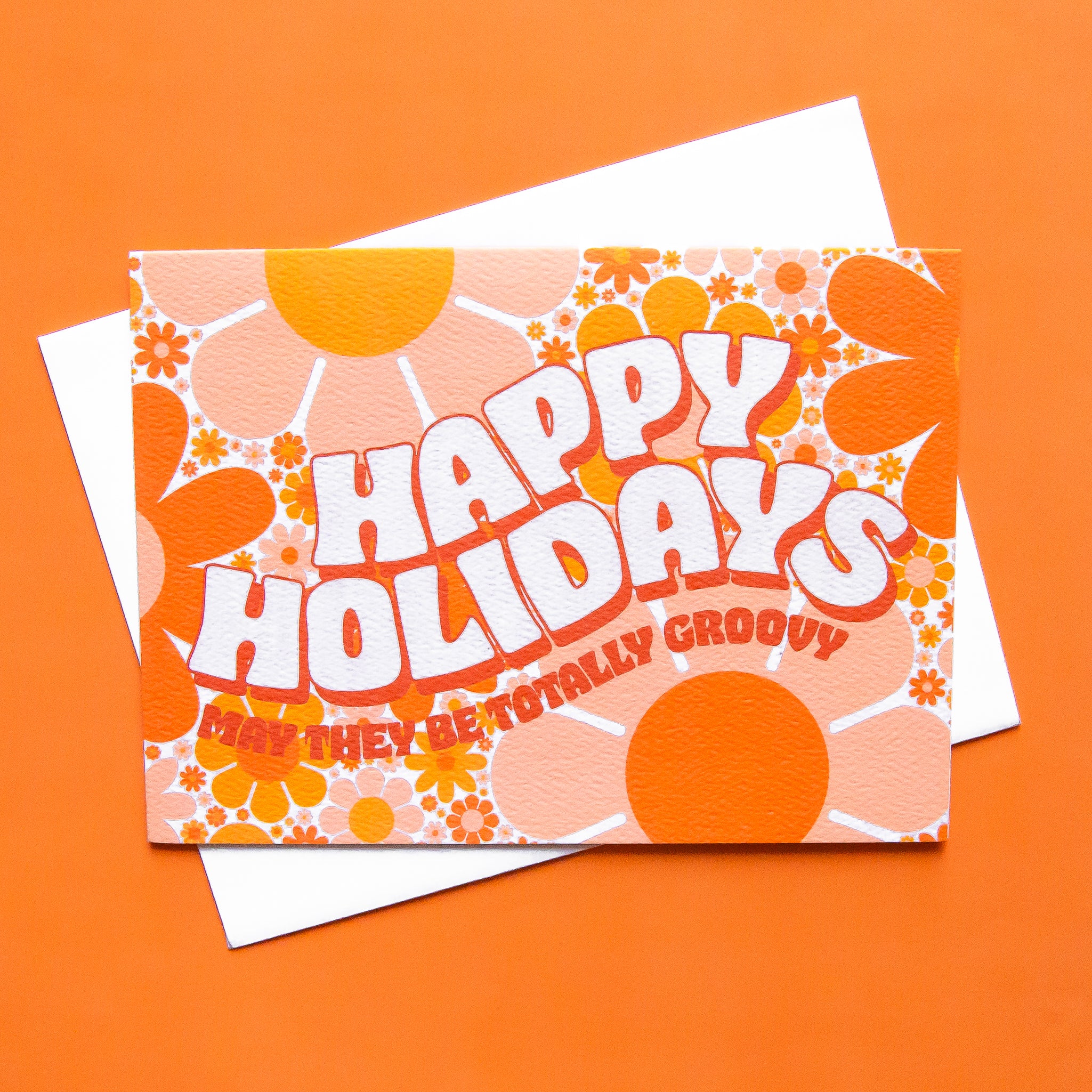 Card filled with a funky 70’s design made up of orange and peach daisies. On top of the design is white block lettering with a red shadow that reads ‘Happy Holidays.’ Under that in red block text it reads ‘may they be totally groovy.’ This card is accompanied by a white envelope.