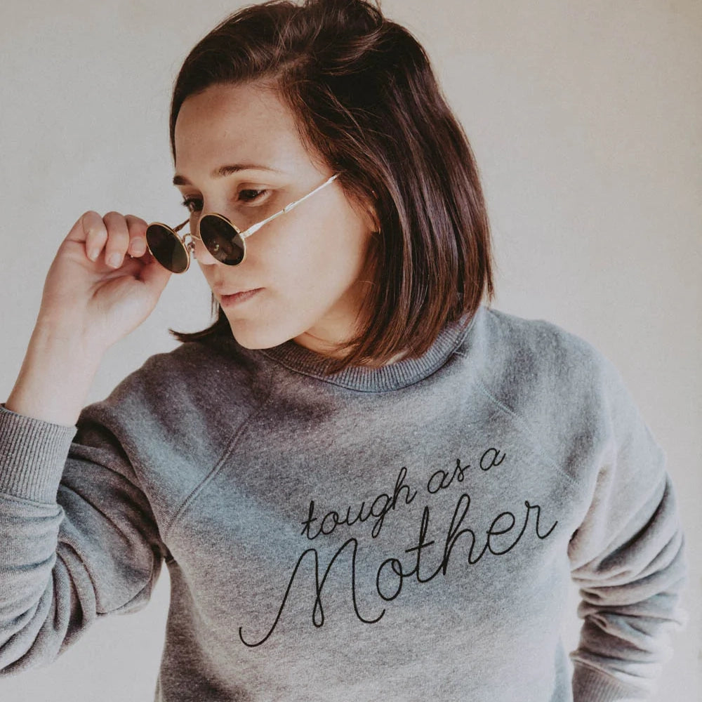 Woman with short brown hair and small round sunglasses in a heather grey sweatshirt with the phrase &quot;tough as a Mother&quot; printed on the front in black cursive font.