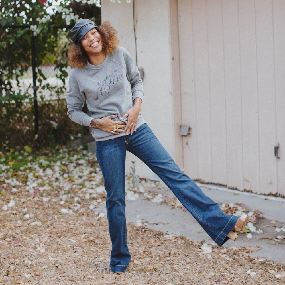 Woman in a heather grey sweatshirt, jeans, and black cap posing with one leg out and both hands on her belly outdoors.