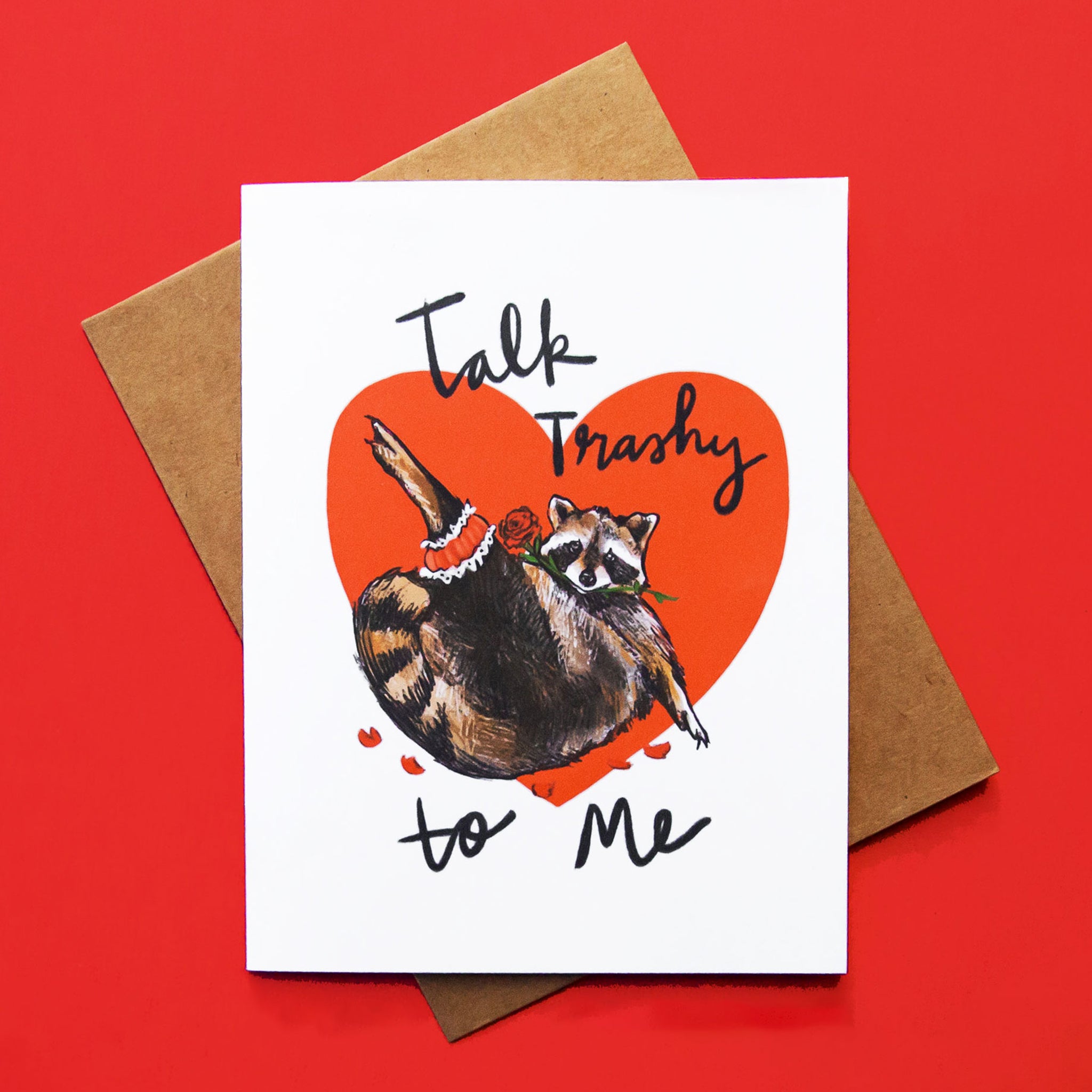 A white card with a red heart and an illustration of a raccoon and text that reads, 'Talk Trashy to me'. 