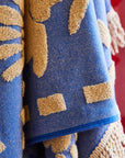 A blue and cream floral pattern bath towel with fringe detailing on two ends. 