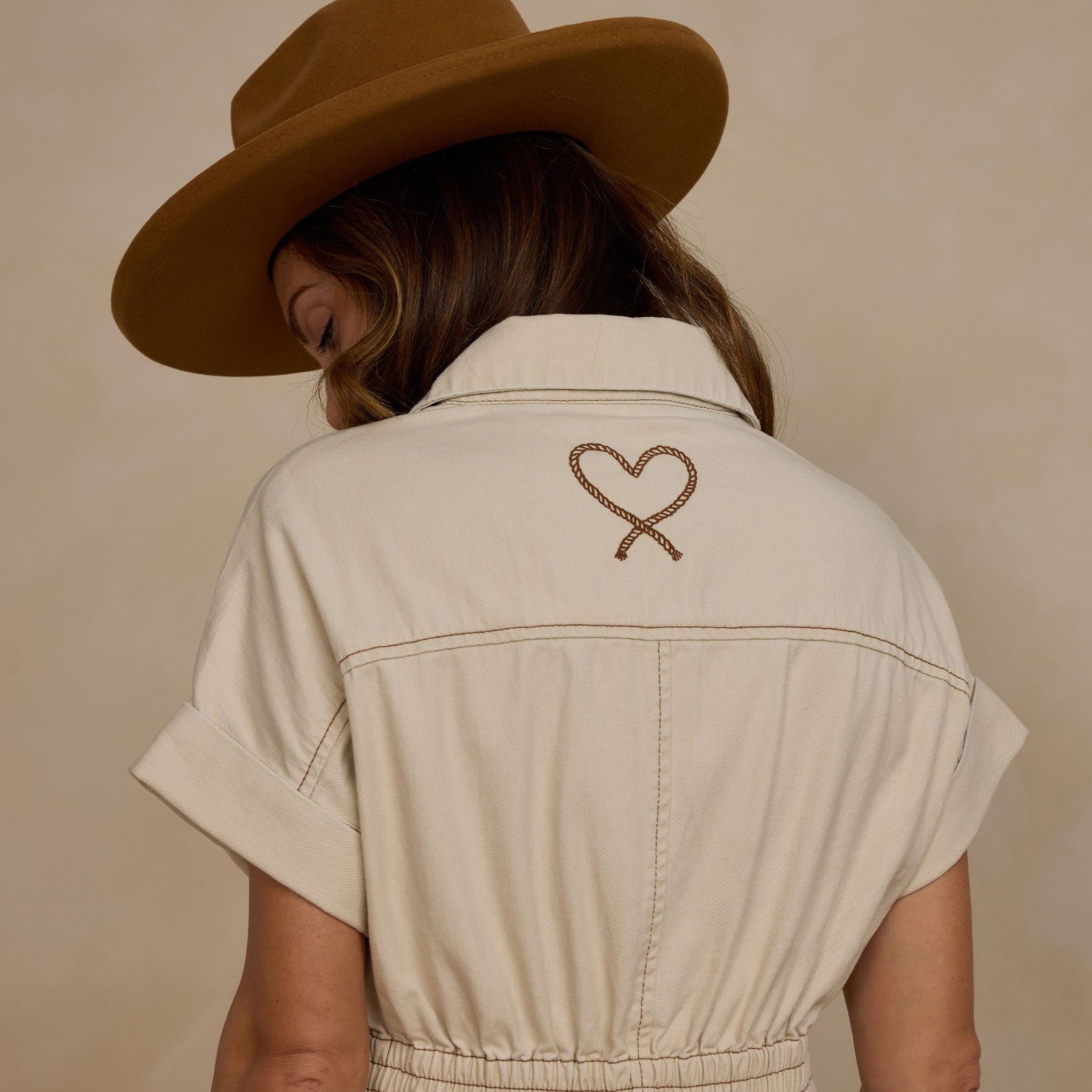 A natural colored short sleeve jumpsuit with a cinched waist and front pockets as well as a stitched rope heart detail on the back.