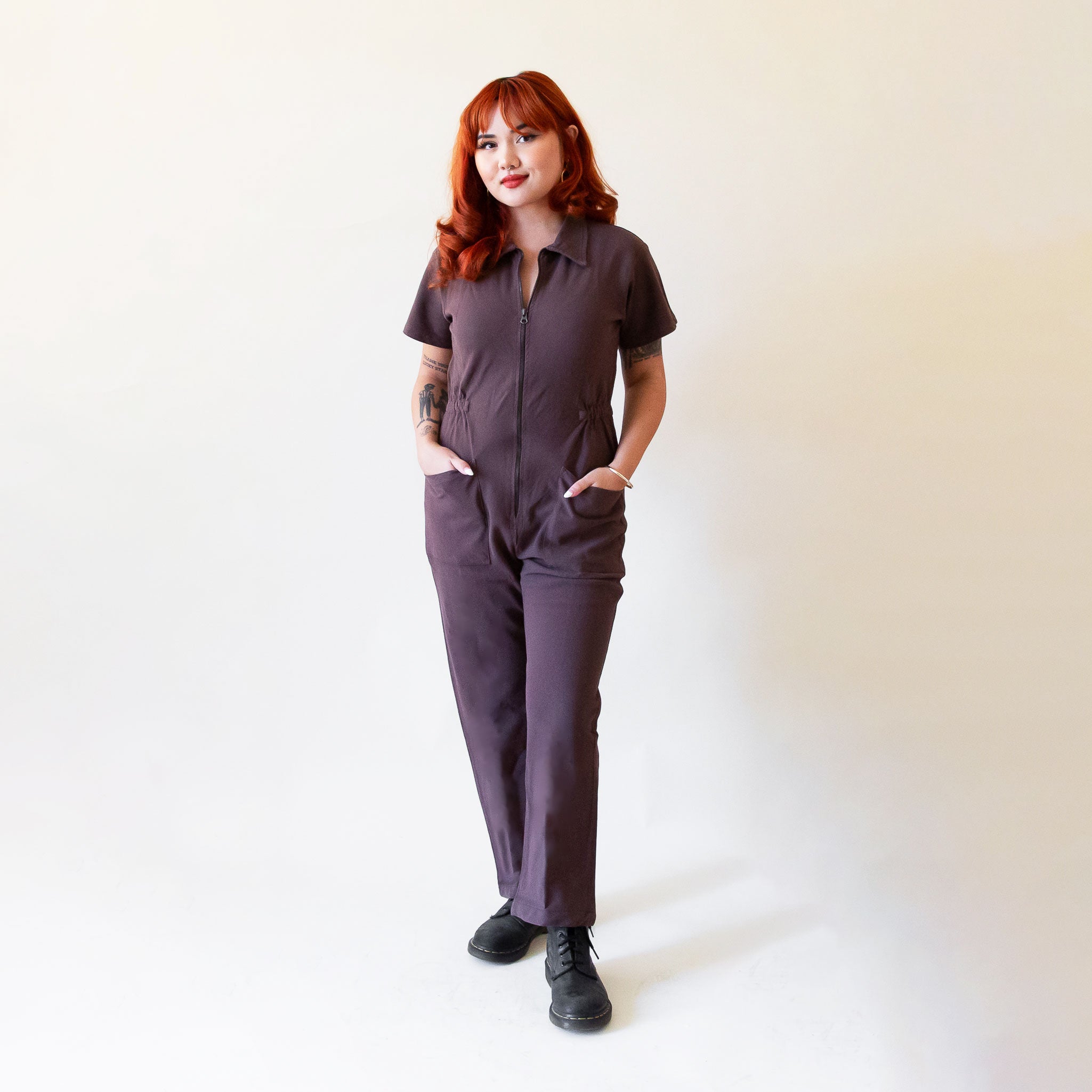 A model standing in front of a bookcase with a chocolate brown utility jumpsuit on with a zipper going up the front, a collar and a cinched waist.