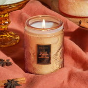 An orange glass, cylinder jar. The glass has a floral texture. On the front is a black sticker with gold text that reads ‘voluspa.’ Below is orange text that reads ‘spiced pumpkin latte.’ Leaning against the left side of jar is a matching glass lid.