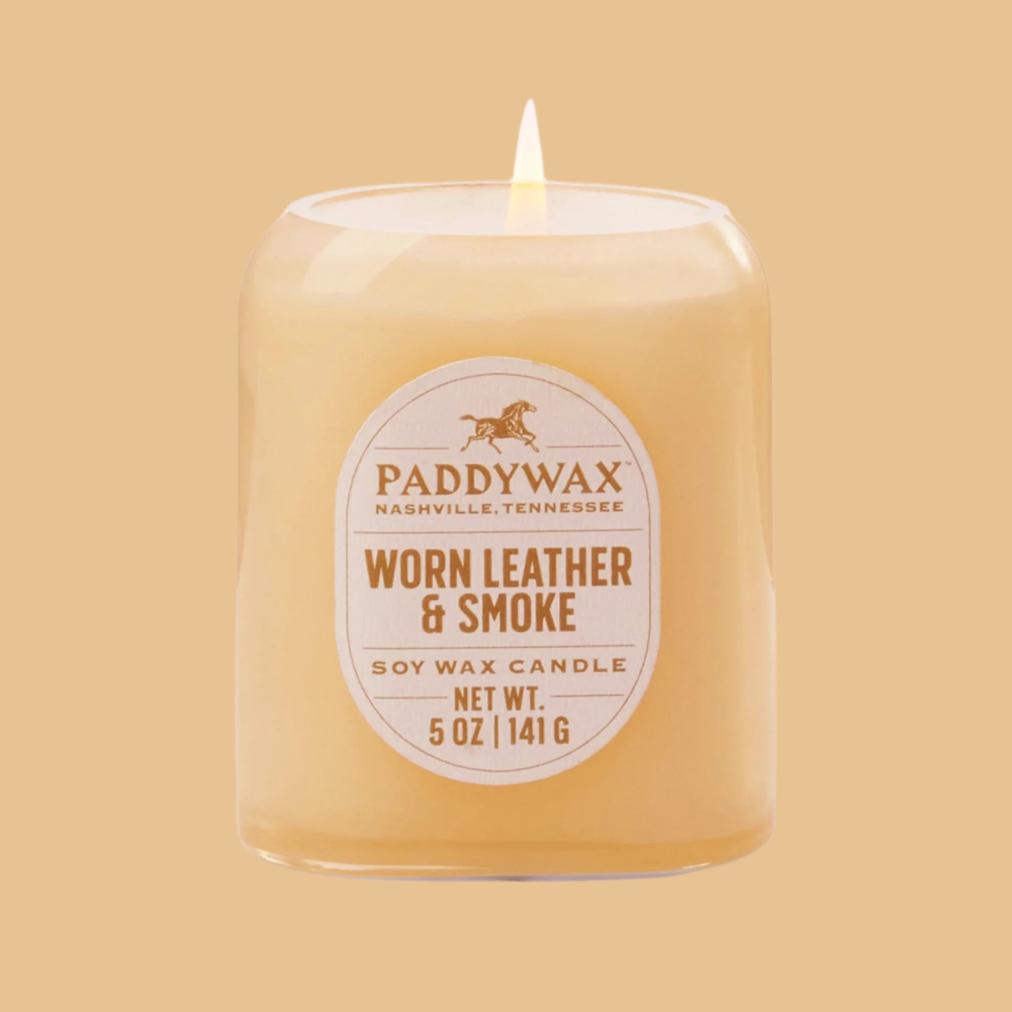 On an orange background is a neutral glass jar candle with an oval label that reads, &quot;Paddywax Worn Leather &amp; Smoke&quot;. 